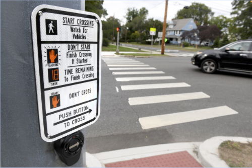 A pedestrian crossing button and sign at a street intersection, with a car blurred in motion, focusing on the button instructions for safely crossing the street.